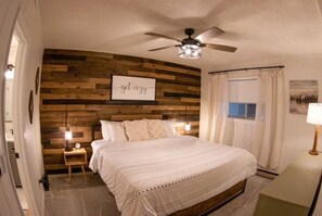 Master bedroom with memory foam king mattress centered on a natural wood accent wall. Matching nightstands with dimmable lamps, with base device changing capabilities. Blackout curtains, ceiling fan with a dimmable light. Full sized dresser.