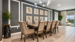 Spacious dining area with a long table, upholstered chairs, modern chandelier, and nature-inspired decor.