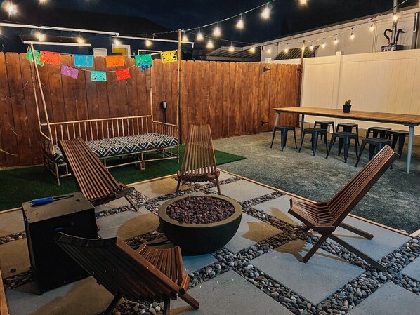 Festive backyard equipped with a propane fire pit, cozy day bed, and large dining table for friends & family to gather. 