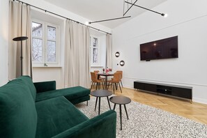 A modern living room featuring a wall-mounted TV, green furniture, and large windows providing a bright atmosphere.