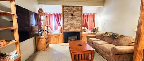The living room features a gas fireplace with a floor-to-ceiling stone hearth situated between two window seats, a couch that can be converted into a queen-size pullout, bookshelves, and a 60” Smart TV.