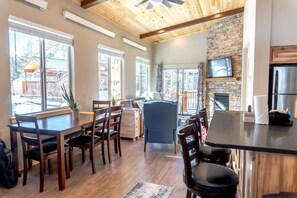 The main living area of the home features an open kitchen/dining/living room area full of natural light and character with high ceilings and exposed wood beams, a gas fireplace in a floor-to-ceiling stone hearth, and windows all around.