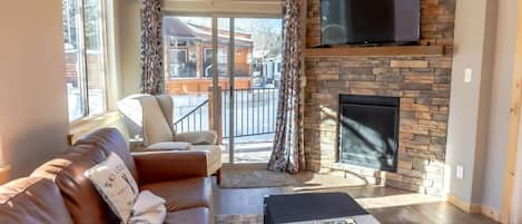 The cozy living area features a leather couch that pulls out to a king-size bed, two reading chairs, a stunning stone fireplace, a 42” flat-screen TV, and access to the outdoor deck from the sliding glass doors.