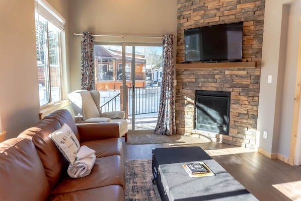 The cozy living area features a leather couch that pulls out to a king-size bed, two reading chairs, a stunning stone fireplace, a 42” flat-screen TV, and access to the outdoor deck from the sliding glass doors.