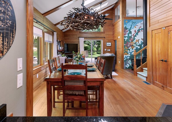 View from kitchen with dining for 6, wood stove on mosaic wall, and light from skylights & doors.