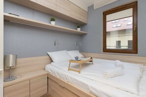 The bedroom has a clean and simple design, with a double bed and light wood furniture. The window provides ample natural light, and the room feels bright and inviting.