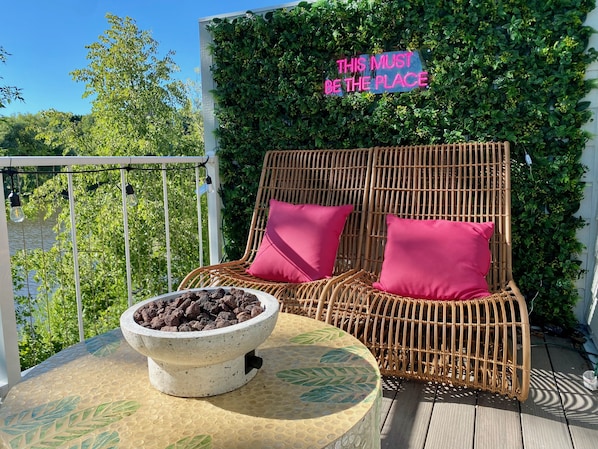 A sunny riverfront deck featuring loungers, a gas-fire table, and a coffee bar. 