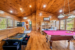 A spacious game room featuring a pool table, foosball table, and a well-lit wooden interior with large windows bringing in natural light.