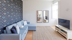Spacious room with the main wall decorated with leaves of different shades of blue. A large TV to enjoy with friends and relatives, A confortable and large resting light-blue sofa and good cushions.