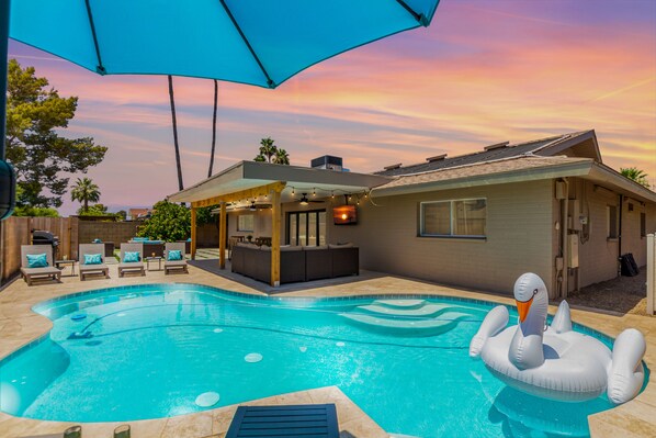 Large backyard with pool, outdoor dining table, outdoor lounging area, putting green, BBQ, cornhole, sun loungers, and fire pit area.