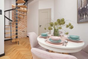 A cozy dining area with pastel pink chairs, a round table set with elegant dishes, and a spiral staircase in the background.

