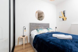 A comfortable bed with a navy blue throw and light-colored pillows. A minimalist lamp sits on a small wooden table beside it.