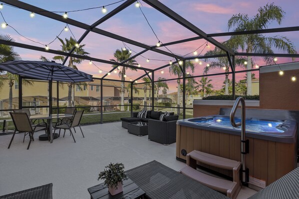 Our home features a cozy outdoor patio with a hot tub, wicker seating, and a dining table under a striped umbrella. String lights create a warm ambiance, and palm trees and neighboring homes are visible in the background, enhancing the tropical