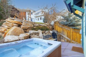 Vacation rental private hot tub on the back deck surrounded by a stone wall, fence, trees, and homes in the distance.