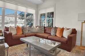 The living room featuring a brown leather sectional, neutral rug, throw pillows, large windows, and a grey coffee table.