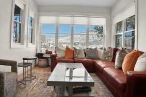 The living room featuring a brown leather sectional, neutral rug, throw pillows, large windows, and a grey coffee table.