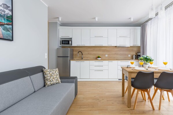 A bright living area combining a cozy sofa, a dining table, and a modern kitchenette with white cabinets and wooden accents.
