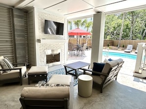 Covered pool-side deck area with comfort seating, a fireplace, and flat-screen television.