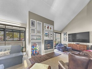 A living room with a large TV, various seating options, a fireplace, and built-in shelves displaying decorative items. A tall window with a scenic view and a toy play area are also visible.