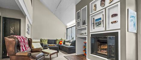 A cozy living room features a leather armchair, an L-shaped sofa, a fireplace with decorated shelves above it, and a brick hearth. Adjacent walls are adorned with artwork and photographs.