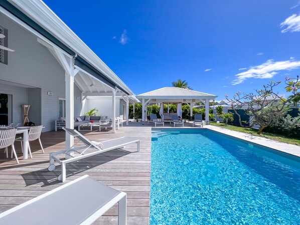 View of the swimming pool, wooden terrace, deckchairs and shaded patio with outdoor seating area