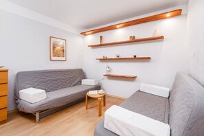 A bright living space featuring a sofa bed, wall-mounted shelves, and warm wooden flooring.
