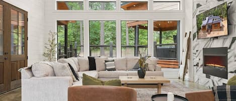 Experience the joy of bonding at Up In Smoke: A modern cabin retreat where your family and friends gather, surrounded by a breathtaking view through floor-to-ceiling windows, a spacious haven adorned with large comfy couch & chairs and cozy throw blankets,
