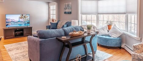 Large living room with couch and love-seat, large TV, wood burning fire place, bay-window with bench and reading nook.