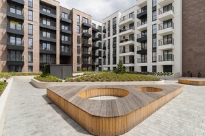 A landscaped courtyard with modern benches and a circular wooden planter in the center. The area is surrounded by contemporary apartment buildings.