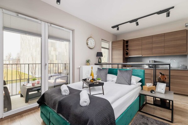 A bright bedroom with a neatly made double bed, green cushions, and matching decor. The room features large windows, allowing plenty of natural light.