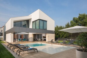 Landschaftsgarten der modernen, neu erbauten Villa mit Pool, umgeben von Liegestühlen, Sonnenschirmen und einem Esstisch im Freien
