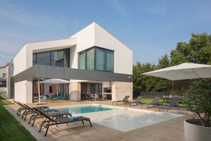 Landscaped yard of the modern newly built villa with pool surrounded by sun loungers, parasols and an outdoor dining table