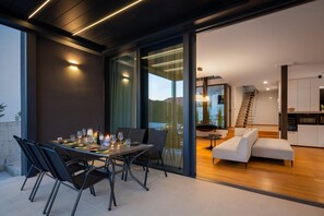 A dining set on the terrace with electronic pergola system and a view on the interior of the Villa with living room and kitchen