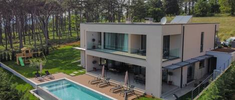 View of the property of the luxurious Villa Paradiso Verde Istria with a 40m2 swimming pool, a sundeck with sunbeds and a shower, and a children's town surrounded by pine forest and greenery