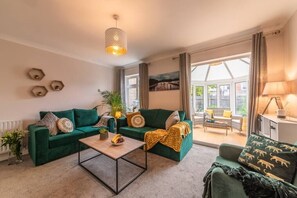 Living area with sofa bed, 2 seater sofa and chair in plush green. There is a door leading to the conservatory and garden. TV present on opposite wall. 