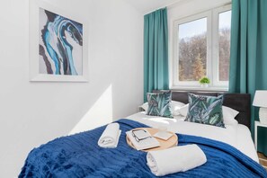 A cozy bedroom featuring a comfortable double bed with blue bedding and green curtains framing the window.
