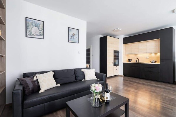 A modern living room with a black sofa, coffee table, and an open view of the kitchen area.
