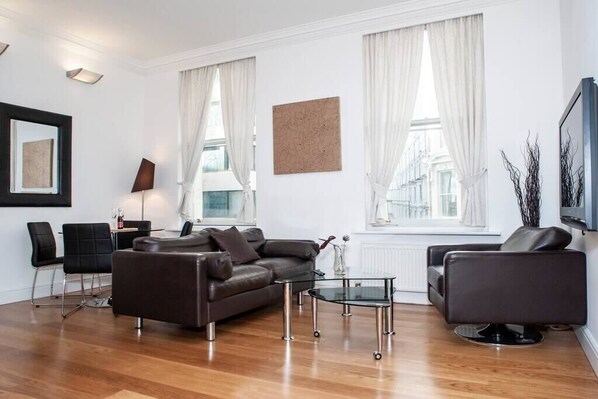 The spacious, bright living room with high ceilings creates an open, airy atmosphere. It's furnished with a comfortable sofa and armchair, perfect for lounging, and a sleek glass round table with chairs, providing a stylish and functional space.