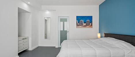 A minimalist bedroom with a white bed, a blue chair, and a beach painting.