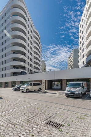 An exterior shot of a modern apartment building with a curved facade, balconies, and a clean paved walkway in the foreground. The sky is clear and blue.
