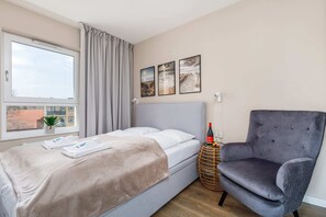 A spacious bedroom with a neatly made double bed, soft gray and white bedding, and a comfortable armchair in the corner. The room is well-lit thanks to a large window.