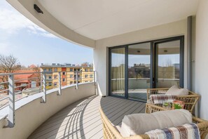 A curved balcony with outdoor seating, including a small round table and cushioned chairs. The balcony railing provides an open view of the surrounding cityscape.