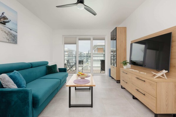 Elegant living room with a comfortable green corner sofa, a TV, and a wooden cabinet, with large windows allowing natural light to flood in.
