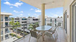 A spacious balcony with a glass-top table and six chairs overlooks modern apartment buildings and trees under a blue sky with scattered clouds.