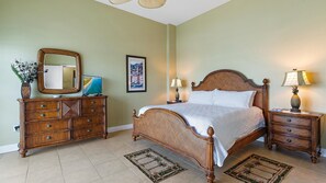 A bedroom with a wooden bed, two nightstands with lamps, a dresser with a mirror, green walls, and a framed picture. There are two small rugs on the tiled floor.