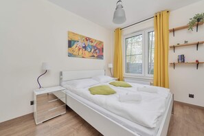 A neat and airy bedroom with a double bed, white bedding, and green accent pillows, along with a colorful wall painting.