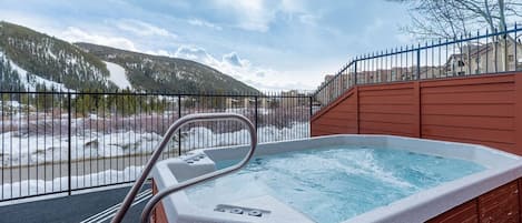 Outdoor hot tub with water jets running, situated on a fenced deck overlooking a snowy mountainous landscape.