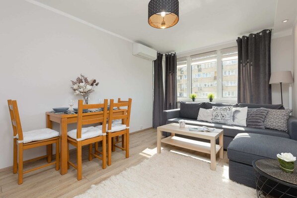 A dining table with wooden chairs, with a lounge area featuring a sofa and a large window letting in natural light in the background.