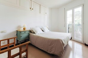 This bedroom is one of three in the townhouse. It features a double bed with soft linens, two bedside tables with lamps, and a small wooden chair. The room is bright and airy with large windows leading to a balcony. The tiled floors are clean and polished