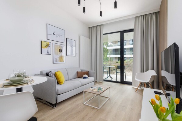 A modern living room with a sleek grey sofa, a coffee table, and a large flat-screen TV. The room is bright, with natural light coming in through floor-to-ceiling windows.